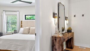 Une chambre à coucher avec un lit, une console en bois surmontée d’un miroir et une fenêtre avec des stores.
