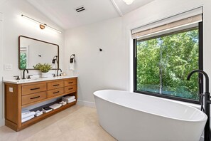 A modern bathroom with a freestanding bathtub, a wooden vanity with drawers, a large mirror, and a window with a view of trees.