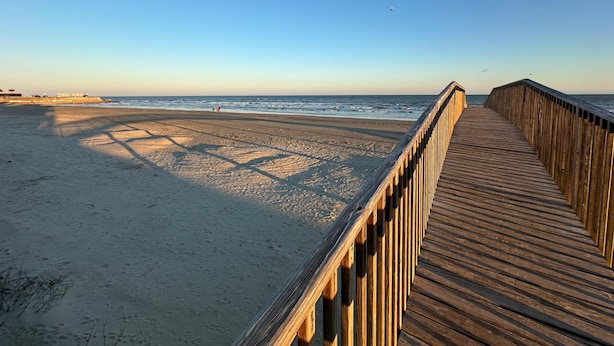 Un paseo marítimo de madera que conduce a una playa arenosa con el océano al fondo.