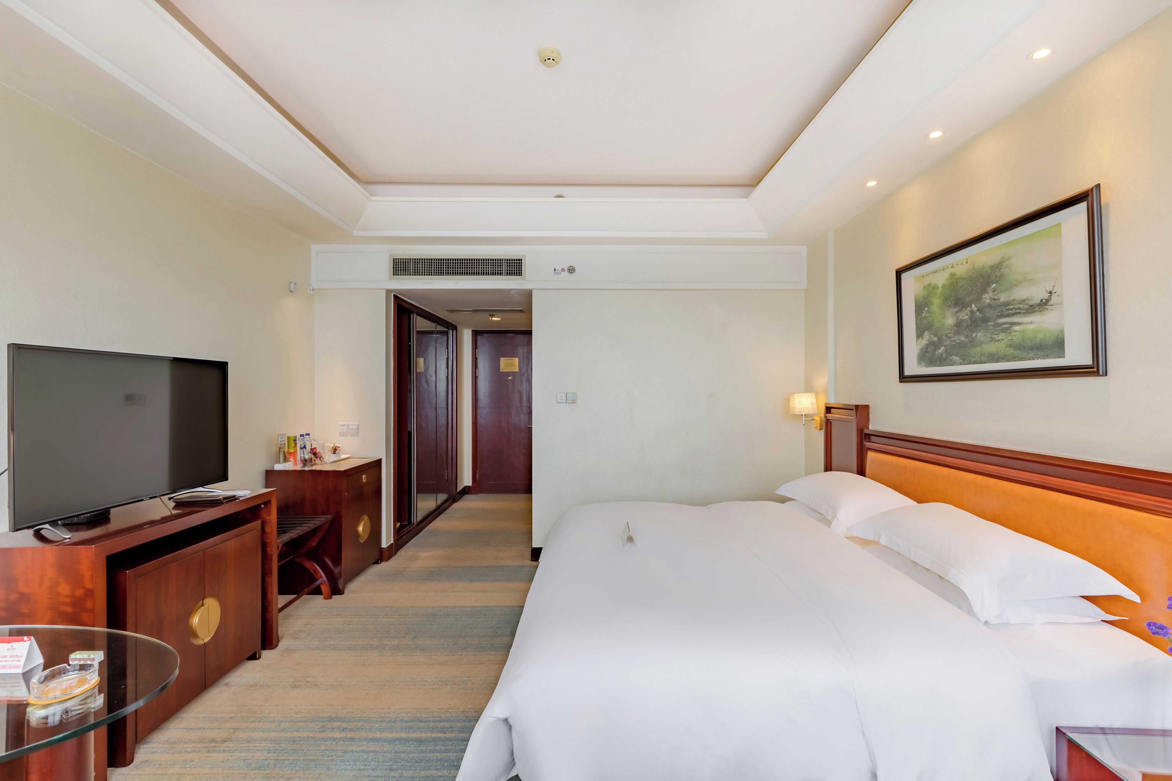 A hotel room with a large bed, a TV, a desk, and a painting on the wall.