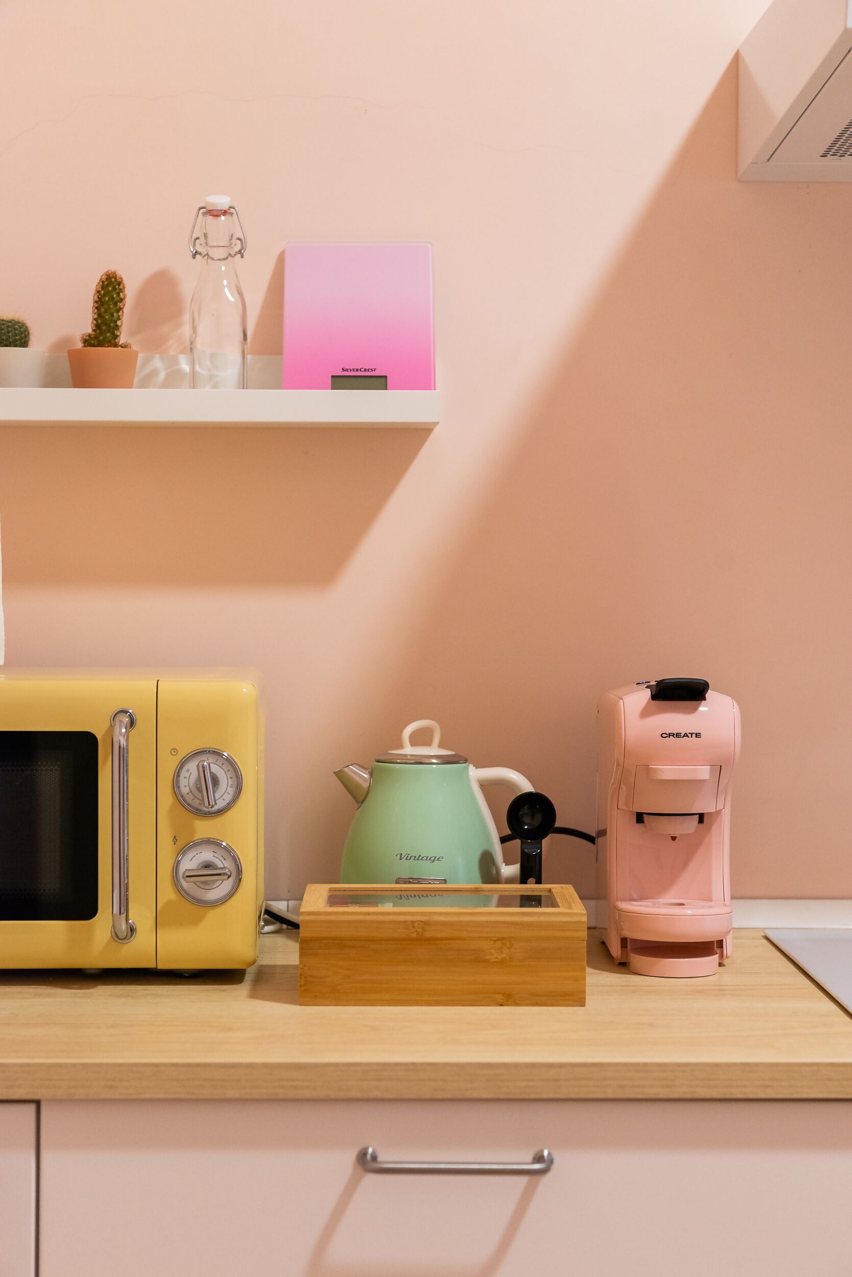 A kitchen with a yellow retro toaster, a green kettle, a pink coffee maker, and a small cactus on a shelf.