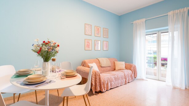 Un salon moderne avec une table à manger ronde, des chaises blanches, un canapé et une grande fenêtre avec des rideaux.