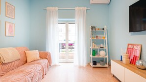 Une pièce avec un mur bleu clair, un canapé blanc recouvert d’un tissu à motifs, une armoire blanche, une étagère contenant divers objets et une fenêtre ornée de rideaux blancs.