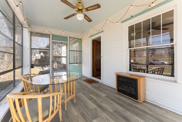 A sunlit room with a glass table, wooden chairs, a ceiling fan, and a fireplace.