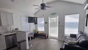 A modern living room with a black leather sofa, a small coffee table, and a TV mounted on the wall. There is a kitchen area with a dishwasher and a small refrigerator. The room has a ceiling fan and large windows with blinds. - Coastal Comfort in Marathon FL2BR Poolside Retreat (Marathon)