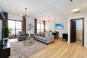 A modern living room with a grey sofa, a coffee table, and a TV. There is a dining area with a table and chairs. The room has large windows and a view of the city.