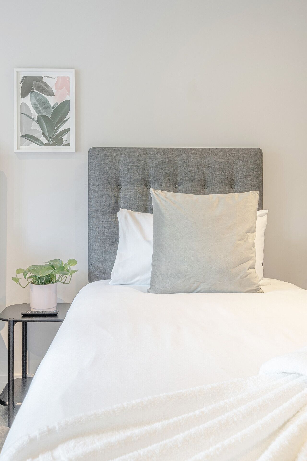 A neatly made bed with a grey upholstered headboard, white pillows, and a white duvet. A framed botanical print hangs on the wall to the left.