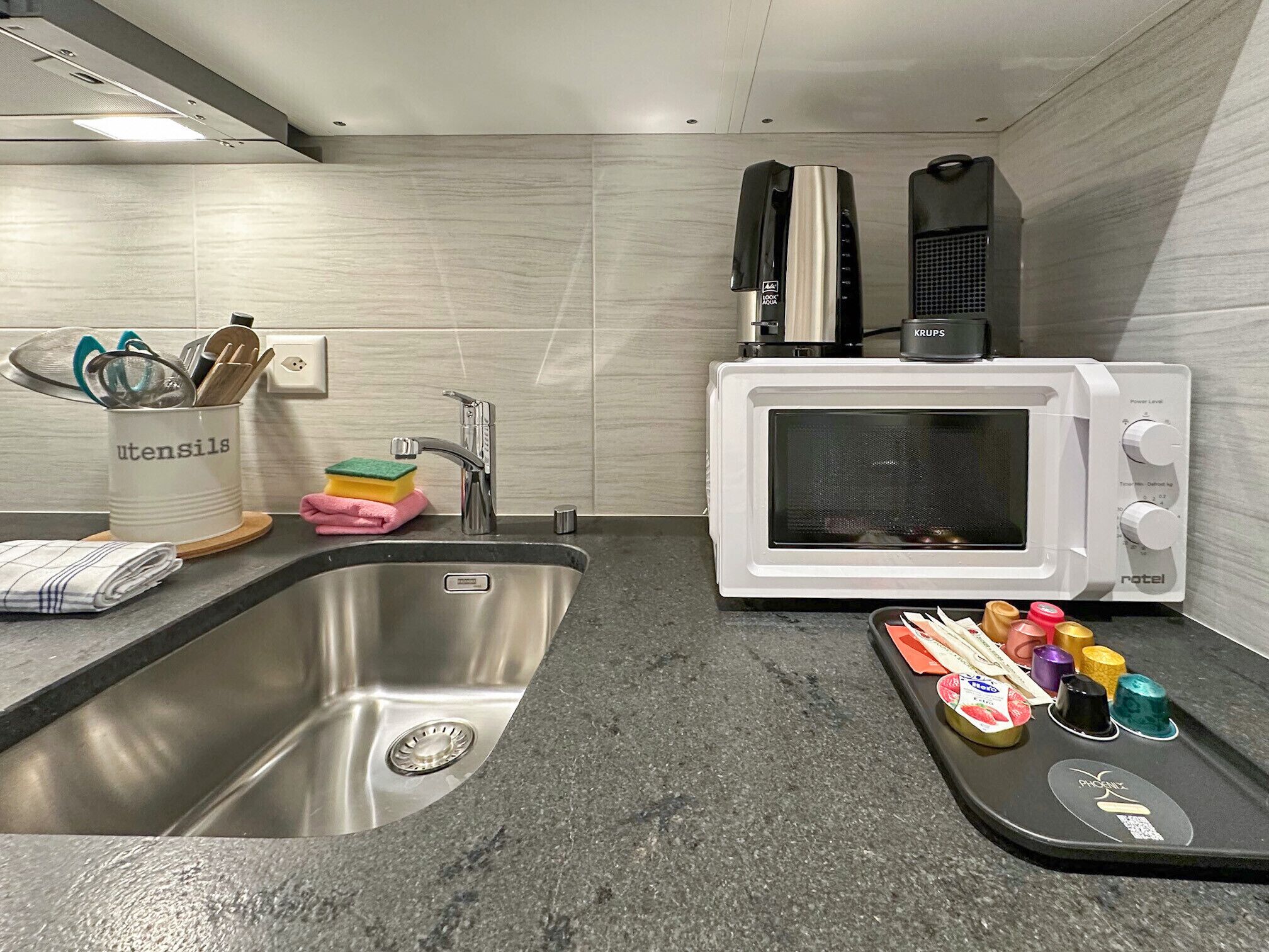 Una cocina moderna con un lavamanos de acero inoxidable, un microondas blanco y una tetera.