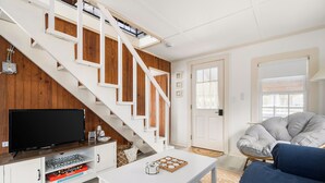 A modern living room with a white staircase, a flat-screen TV, a sofa, and a coffee table. - New! Beach Cottage - 3 min walk to Beach - Pet Friendly! (Dennis)