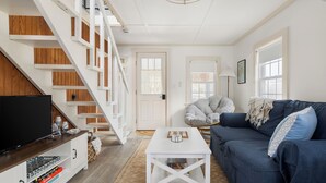 A modern living room with a staircase, a blue sofa, a white coffee table, and a TV on a wooden stand. - New! Beach Cottage - 3 min walk to Beach - Pet Friendly! (Dennis)