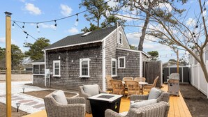 A wooden deck with wicker furniture and a fire pit, surrounded by a house with a dark shingled exterior. - New! Beach Cottage - 3 min walk to Beach - Pet Friendly! (Dennis)