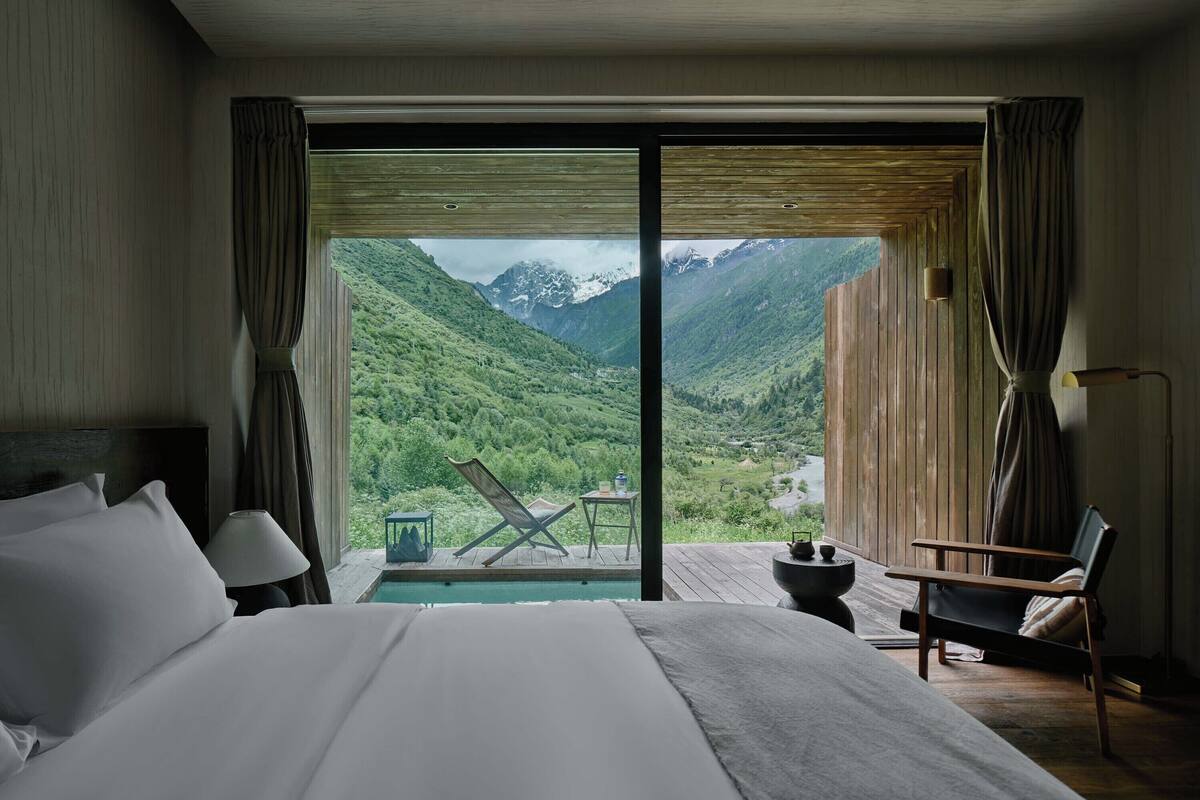 A bedroom with a large bed, a chair, a small table, and a view of a mountainous landscape through a sliding door.