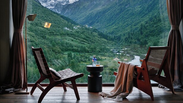 A room with a large window offering a view of a mountainous landscape. There are two wooden chairs and a small table with a bottle and glass. - DAINN Mt Siguniang (Ngawa Autonomous Prefecture)