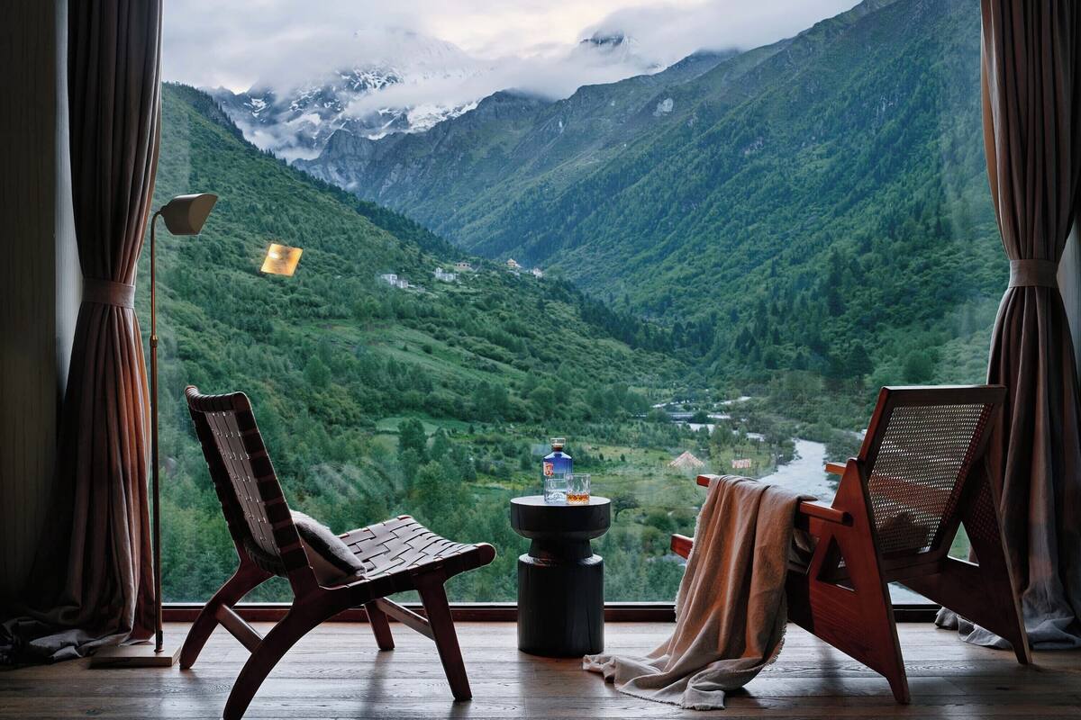 A room with a large window offering a view of a mountainous landscape. There are two wooden chairs and a small table with a bottle and glass.