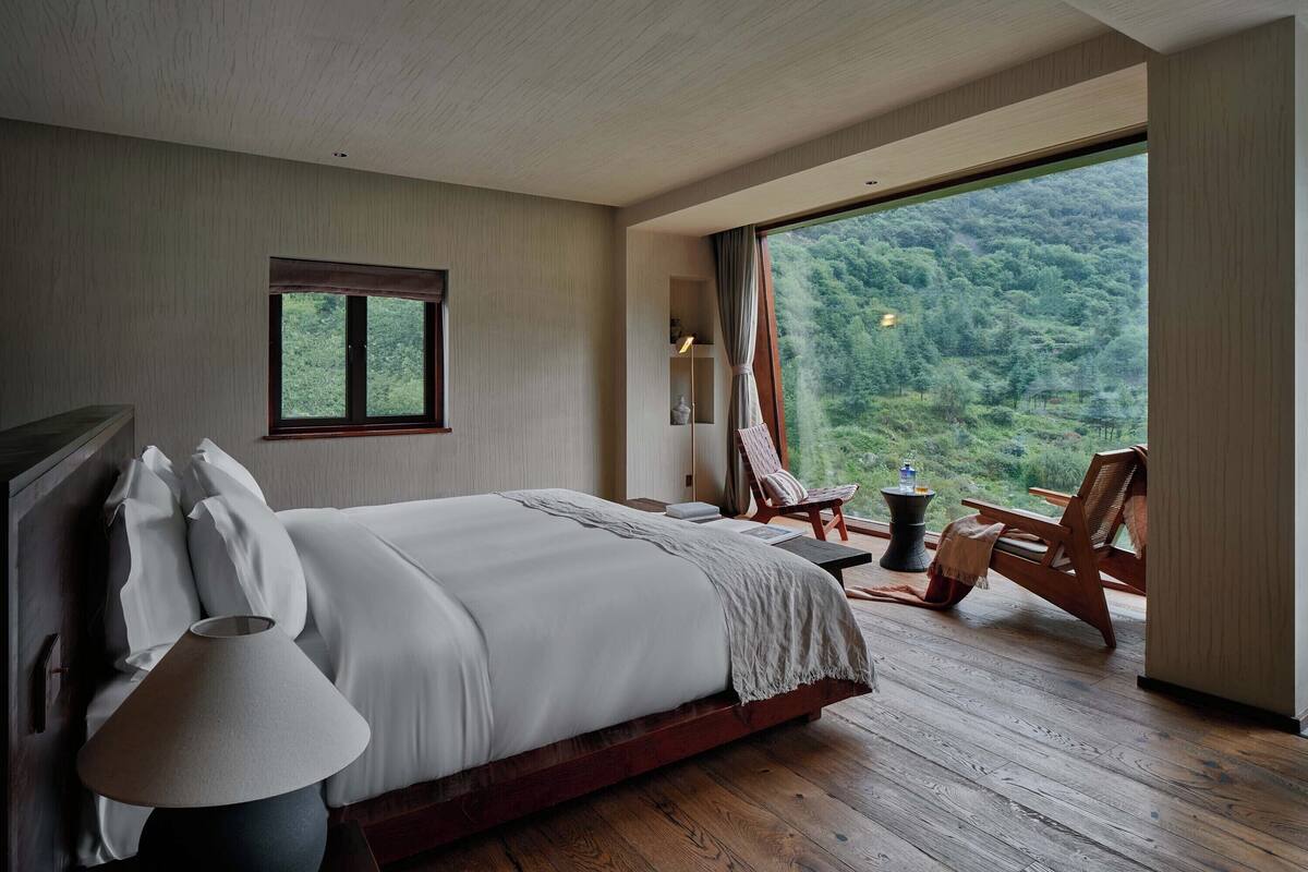 A bedroom with a large bed, a side table, a chair, and a window offering a view of a forested area.