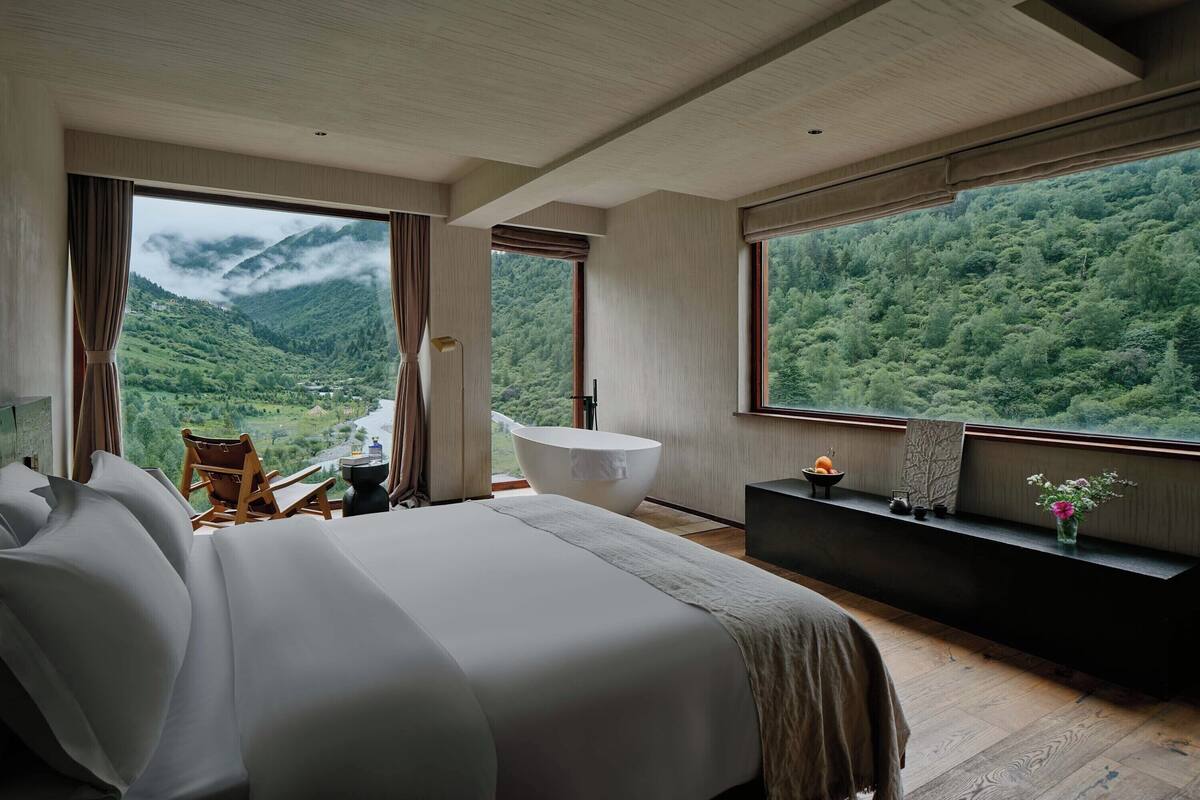 A bedroom with a large bed, a seating area, a bathtub, and a view of a mountainous landscape.
