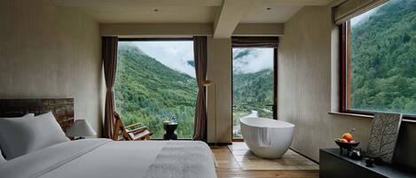 A bedroom with a large bed, a sitting area, a bathtub, and a view of a mountainous landscape.