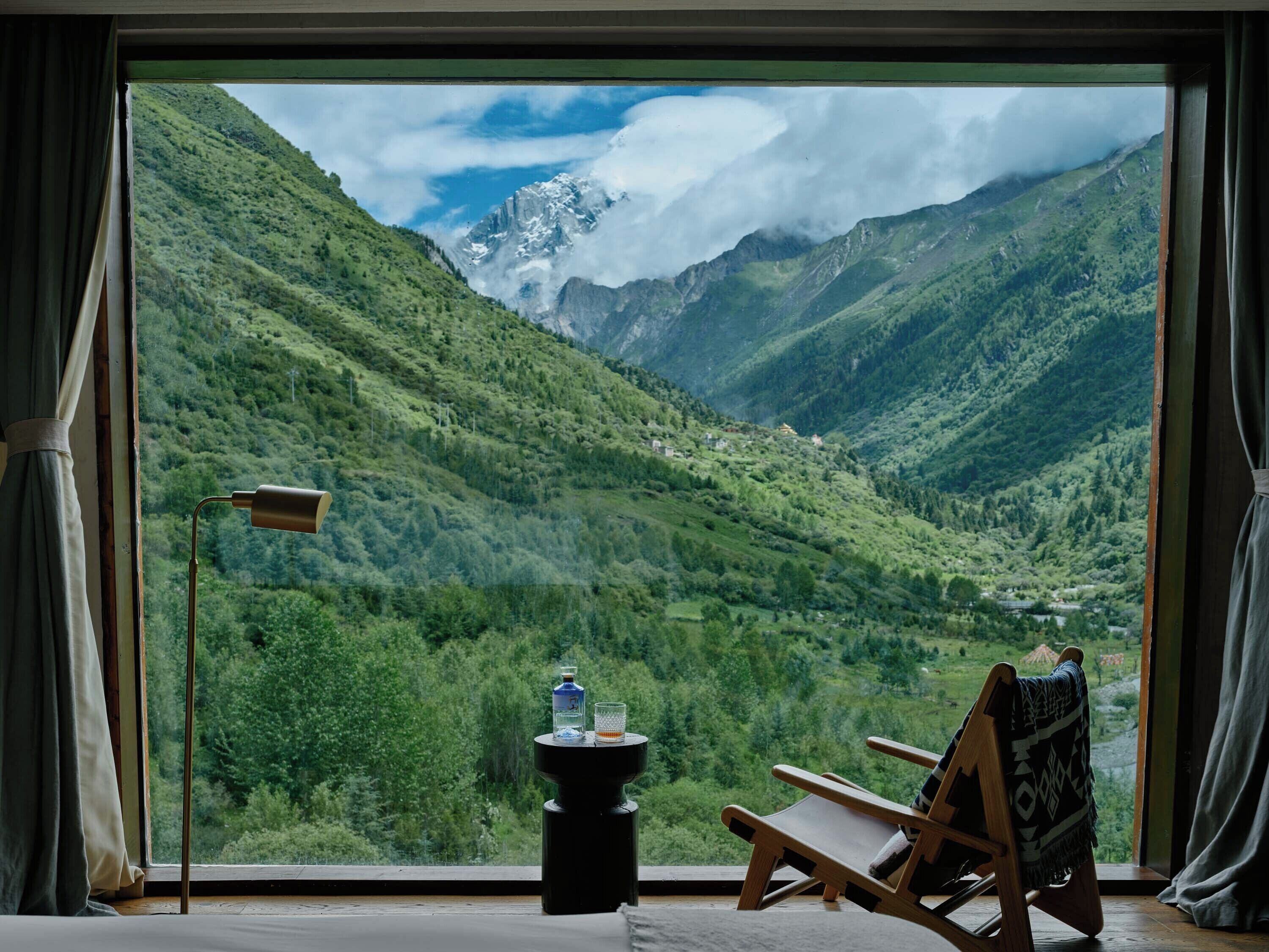 A room with a large window offering a mountain view, a wooden chair, a small table with a bottle and glass, and a lamp.