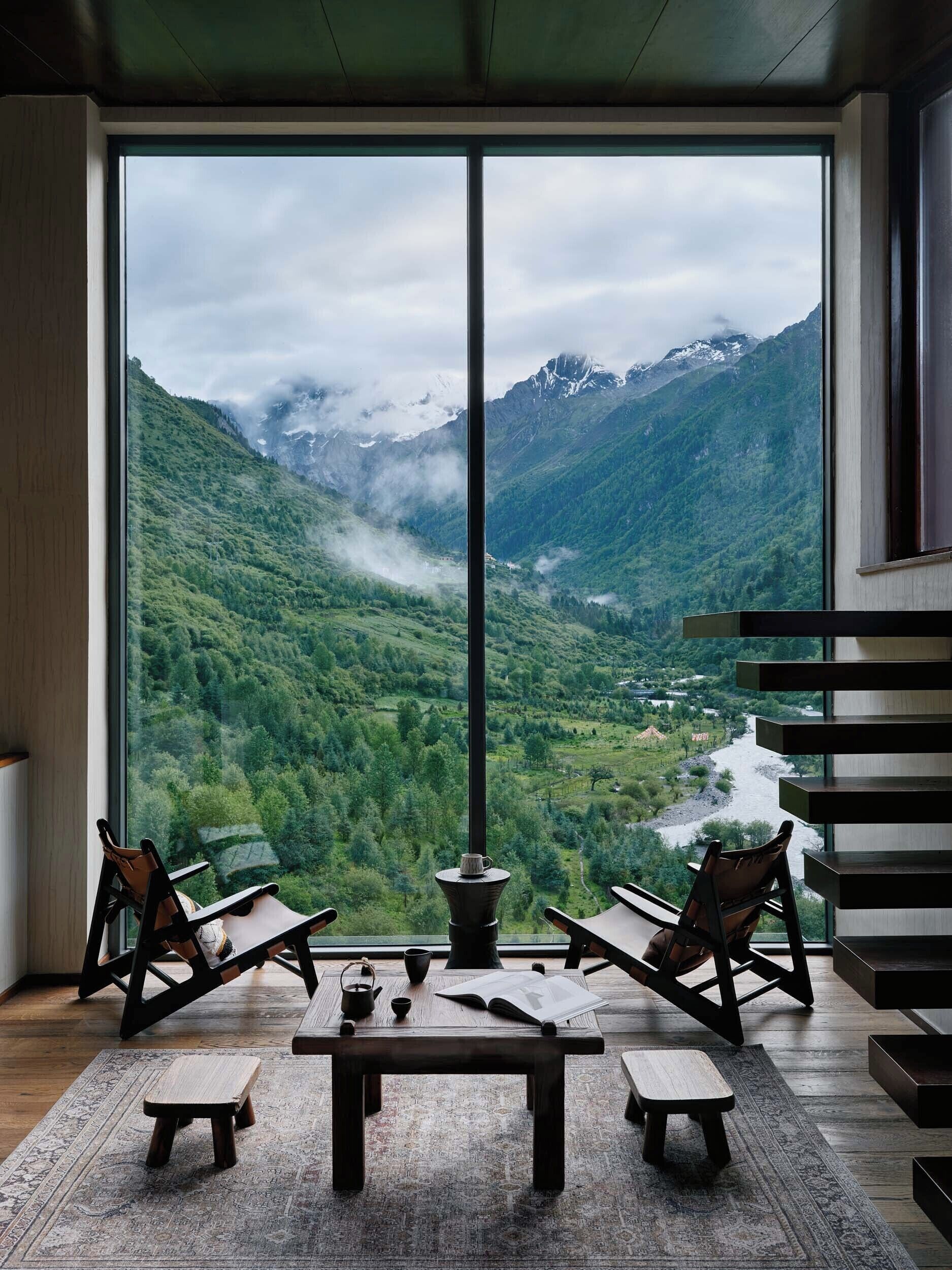 A room with a large window offering a view of a mountainous landscape. There is a wooden table with chairs and a small stool.