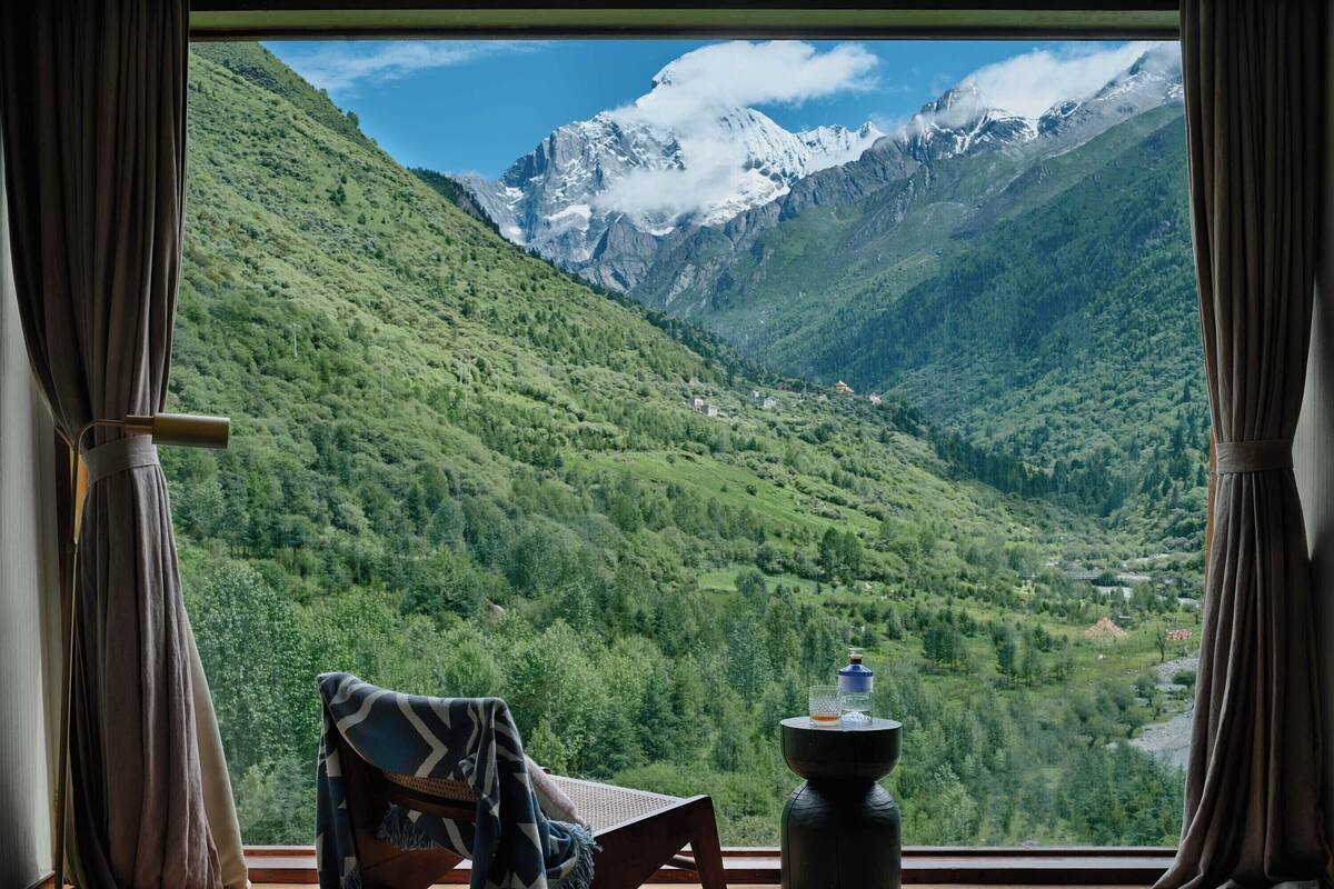 A room with a view of a mountainous landscape, a chair, a table, and a window with curtains.