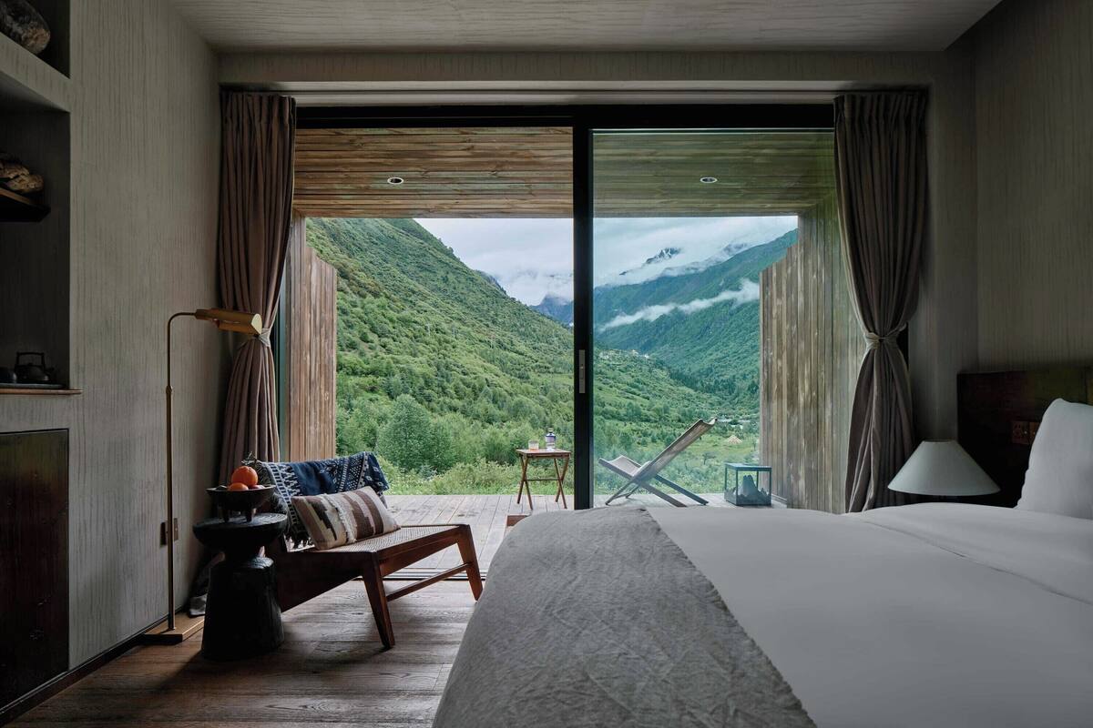 A hotel room with a large bed, a wooden bench, a small table, and a view of a mountainous landscape.