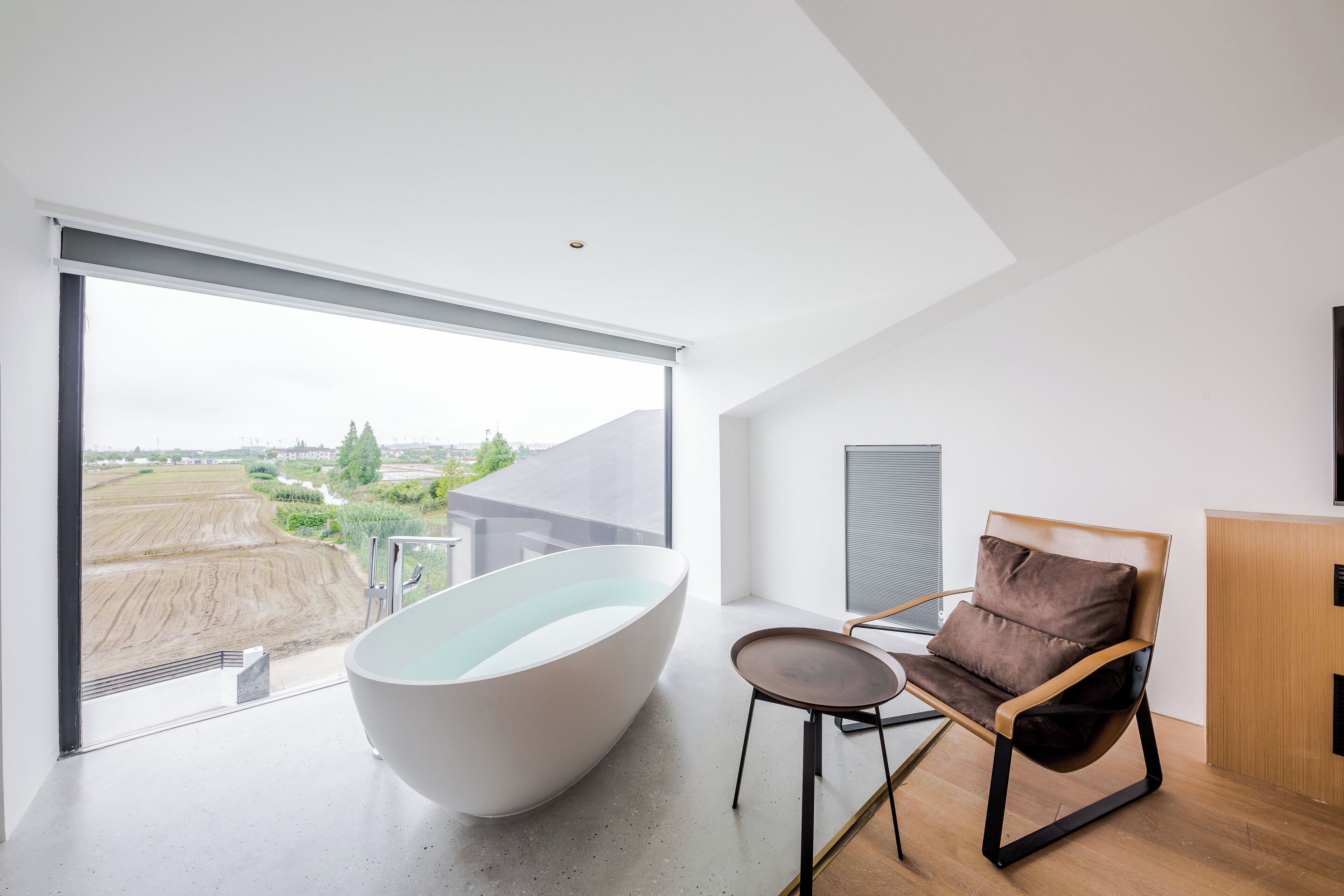 A modern bathroom with a large bathtub, a chair, and a view of a landscape through a large window.