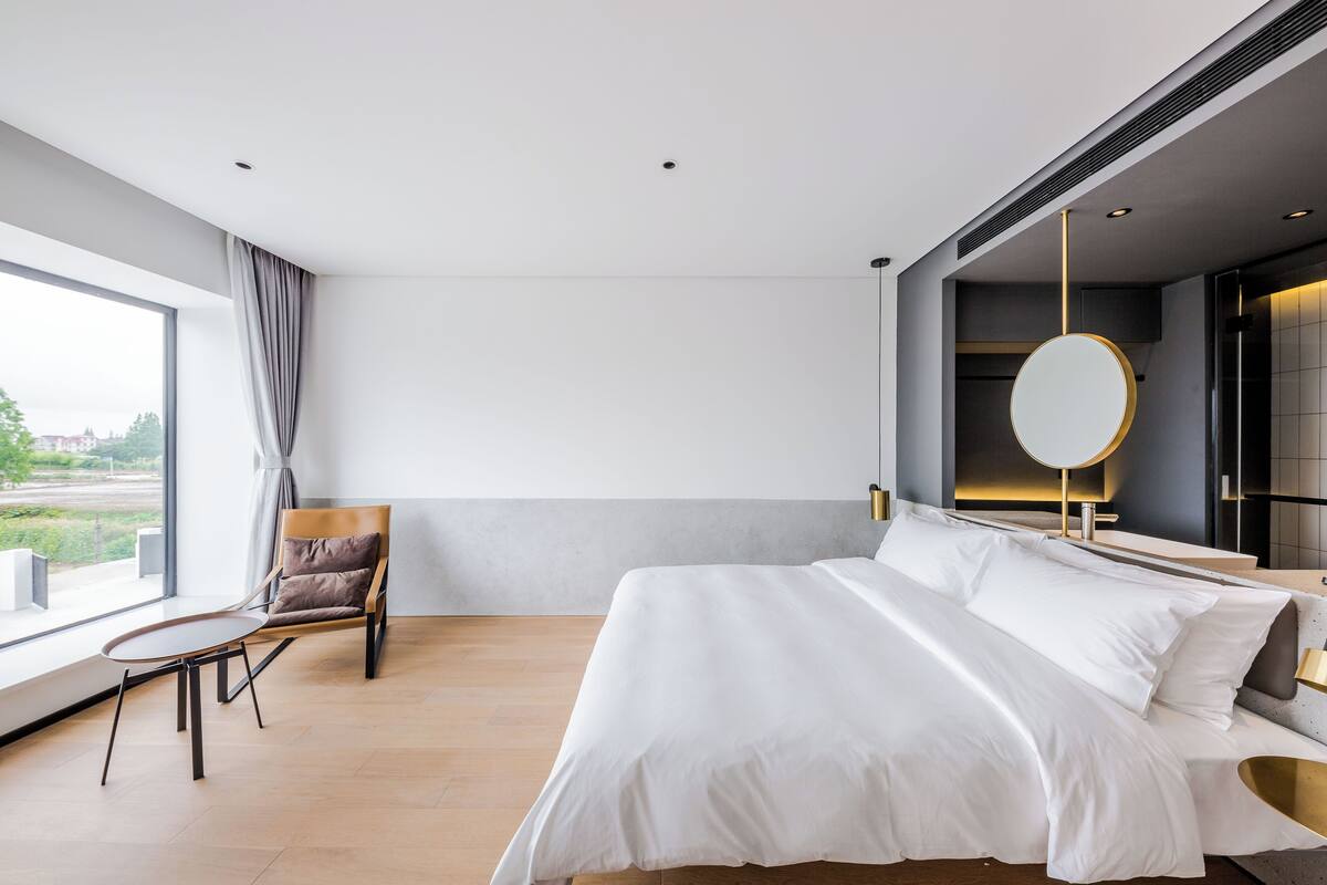 Ein modernes Hotelzimmer mit einem großen Bett, einem Sessel, einem Tisch und Blick ins Freie.