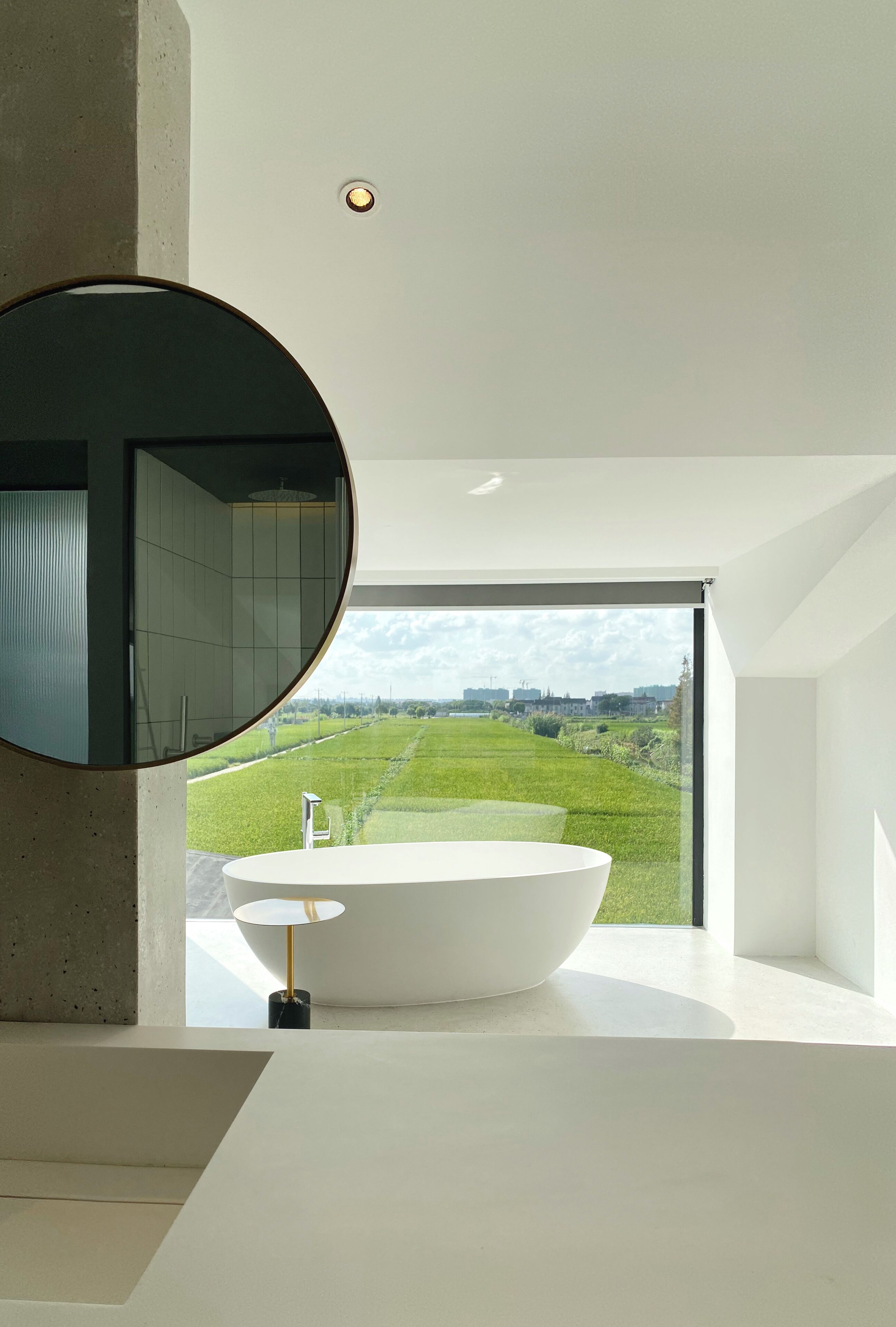 Une salle de bain moderne avec un grand lavabo ovale, un miroir rond et une vue sur un paysage verdoyant par une grande fenêtre.