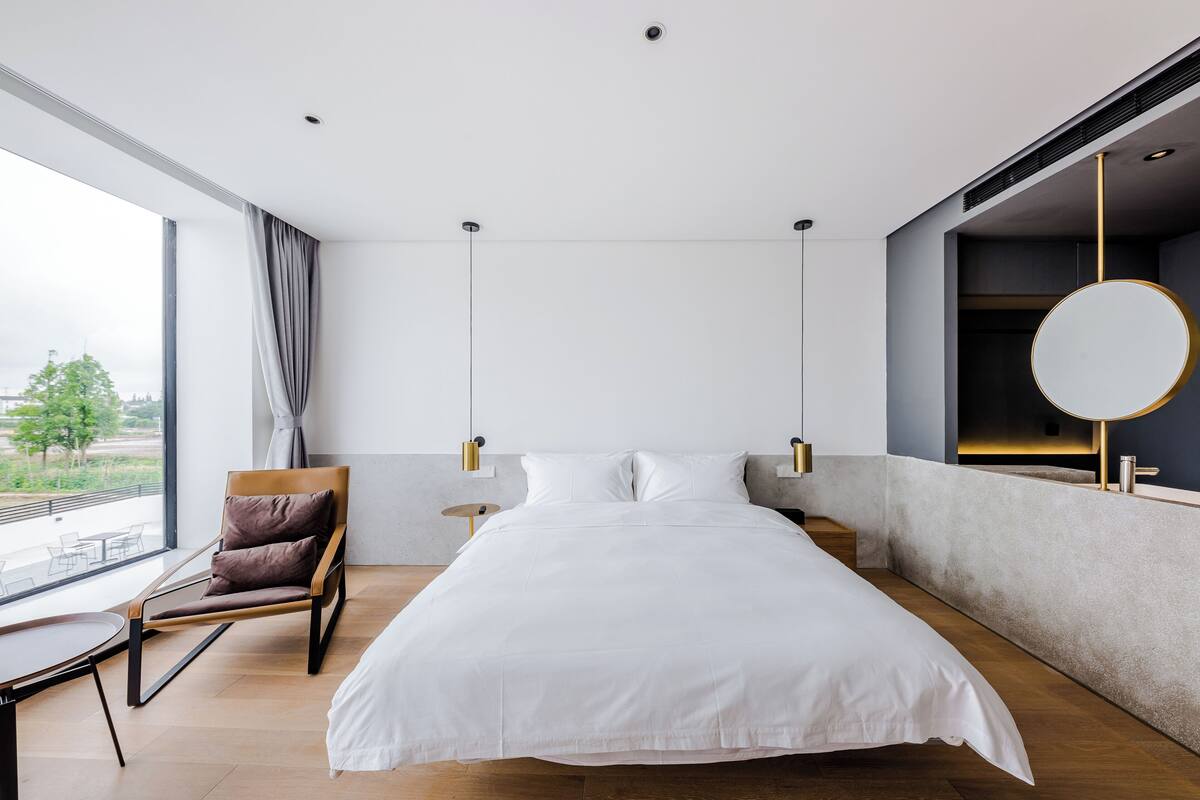 Ein modernes Hotelzimmer mit einem großen Bett, einer Sitzecke mit einem Sessel, einem runden Spiegel und Blick ins Freie.