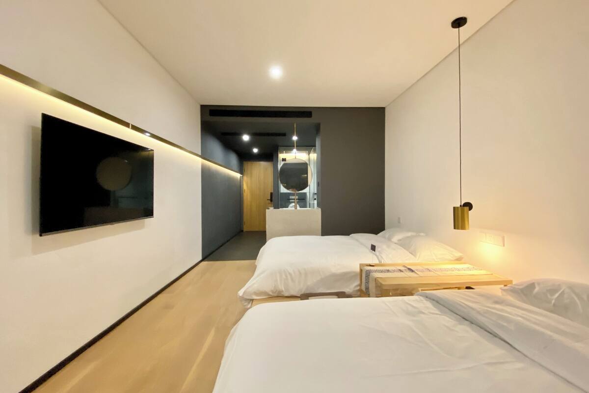 Ein modernes Hotelzimmer mit großem Flachbildfernseher an der Wand, einem Bett mit weißer Bettwäsche, einem Nachttisch und einer hängenden Pendelleuchte.