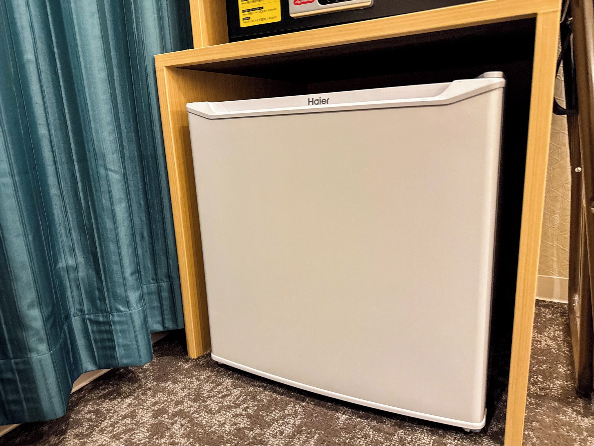 A compact white refrigerator in a wooden cabinet, situated in a room with a patterned carpet and a blue curtain.