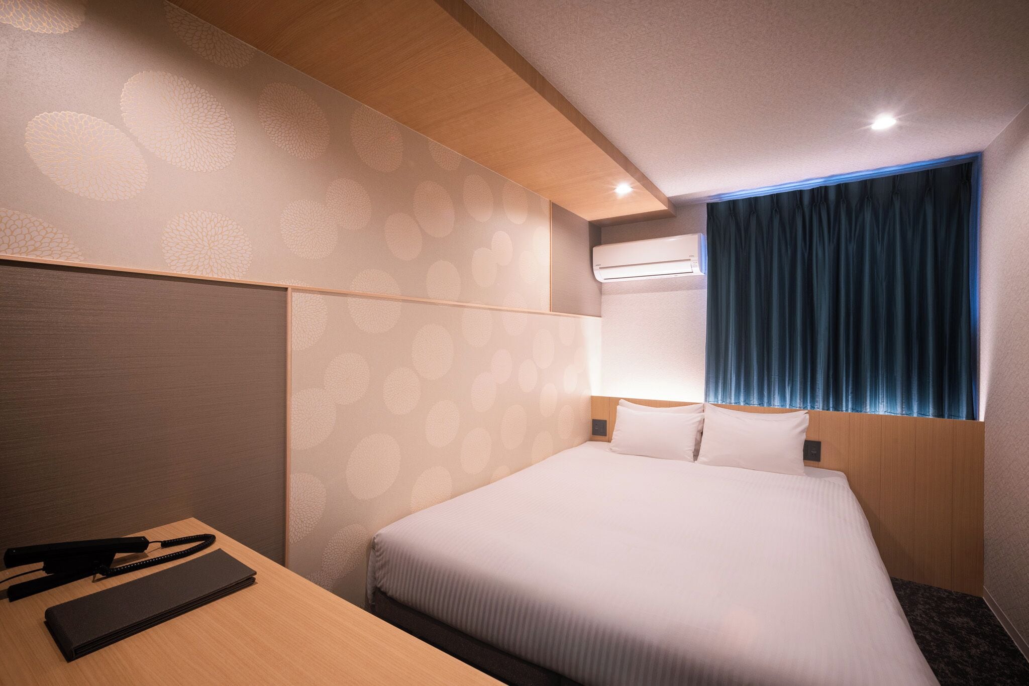 A hotel room with a bed, a desk, a telephone, and an air conditioning unit.