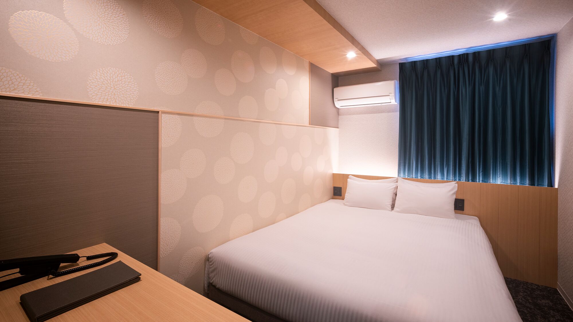 A hotel room with a bed, a desk, a chair, a window with curtains, and an air conditioning unit.