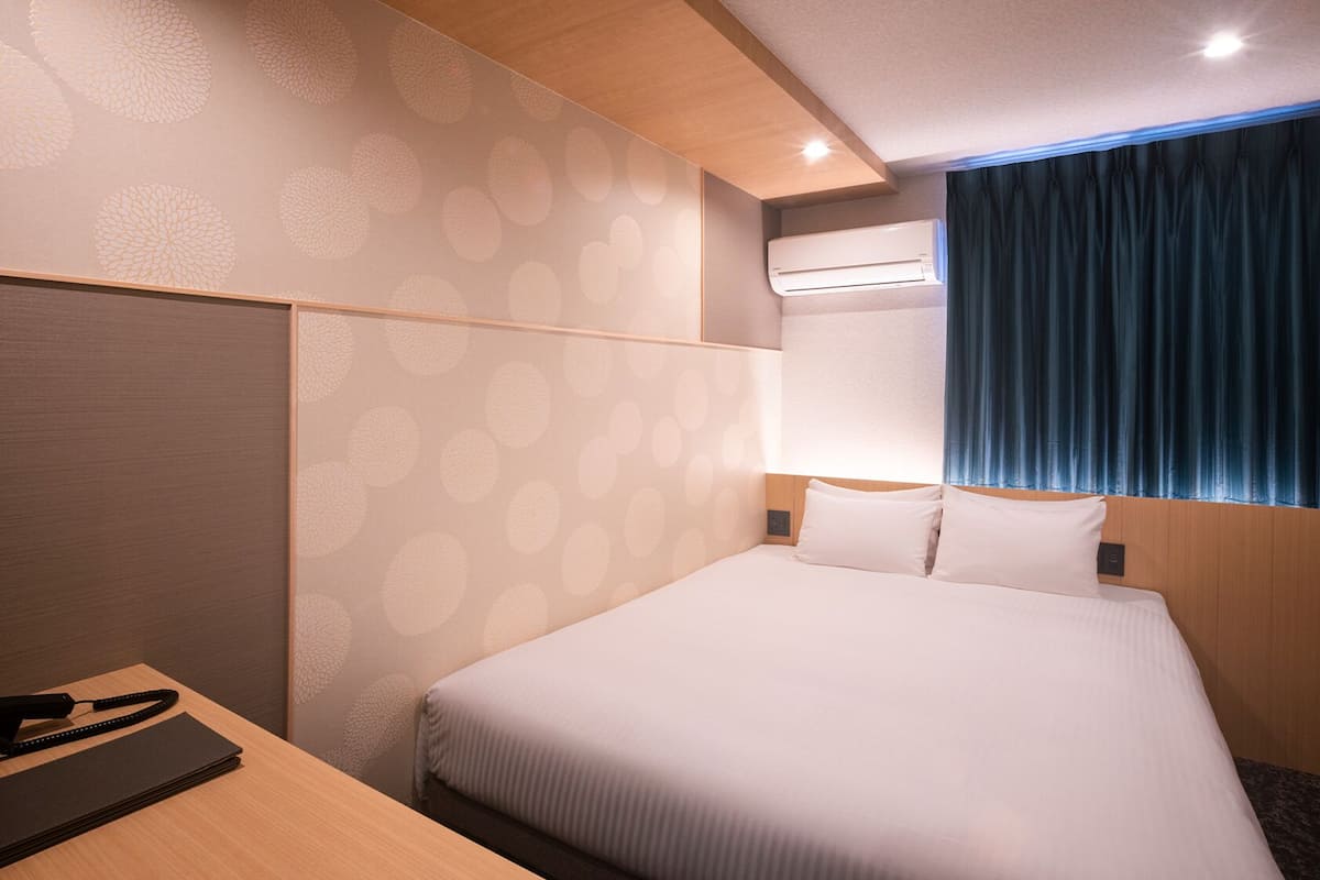 A hotel room with a bed, a desk, a chair, a window with curtains, and an air conditioning unit.