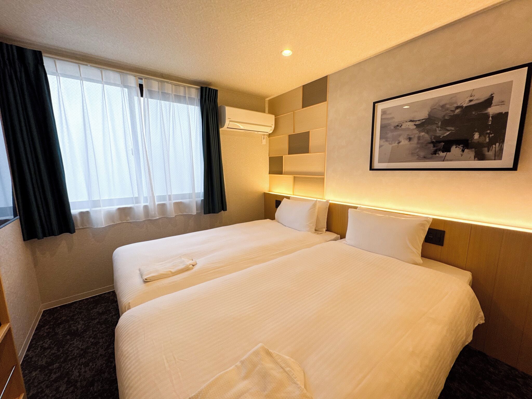 A hotel room with two beds, a large window with sheer curtains, an air conditioning unit, a wall-mounted light, and a framed picture.