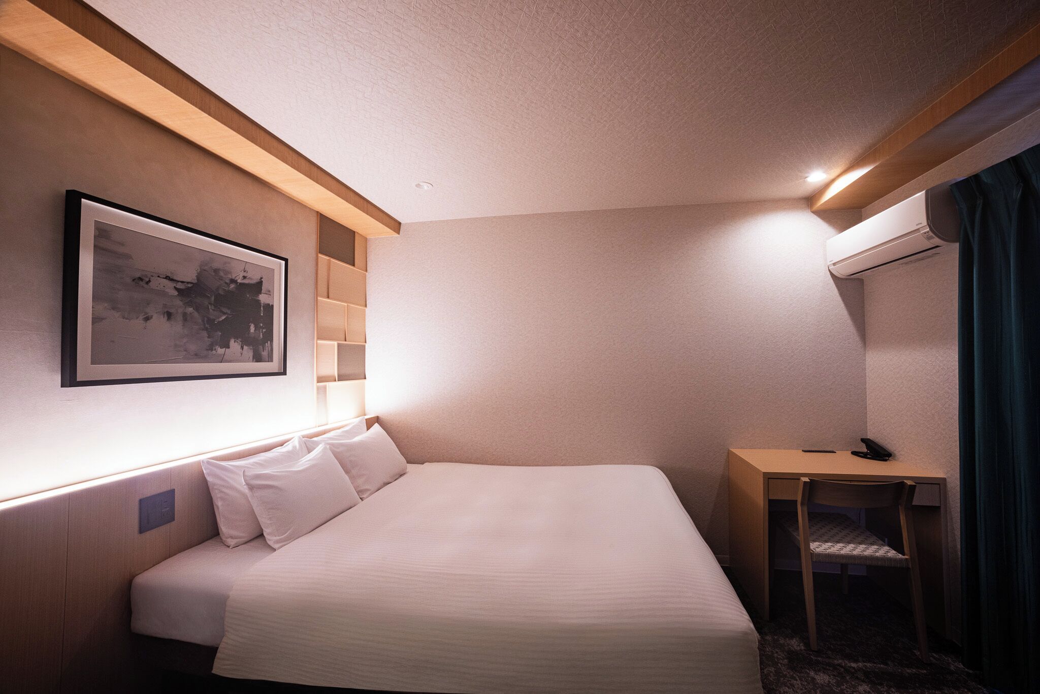 A hotel room with a bed, a desk, a chair, an air conditioner, a framed picture on the wall, and a window with curtains.