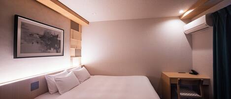 A hotel room with a bed, a desk, a chair, an air conditioner, a framed picture on the wall, and a window with curtains.