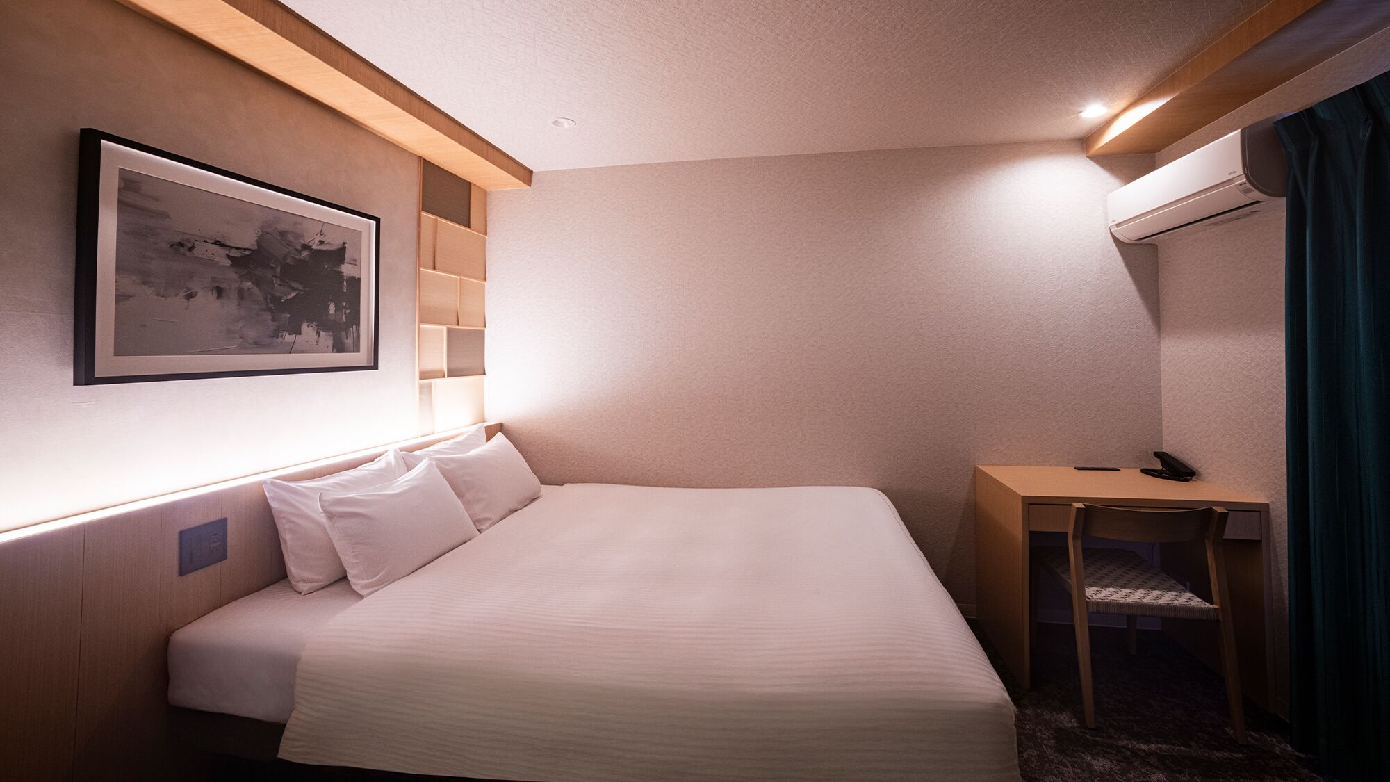 A hotel room with a bed, a desk, a chair, a picture on the wall, and an air conditioning unit.