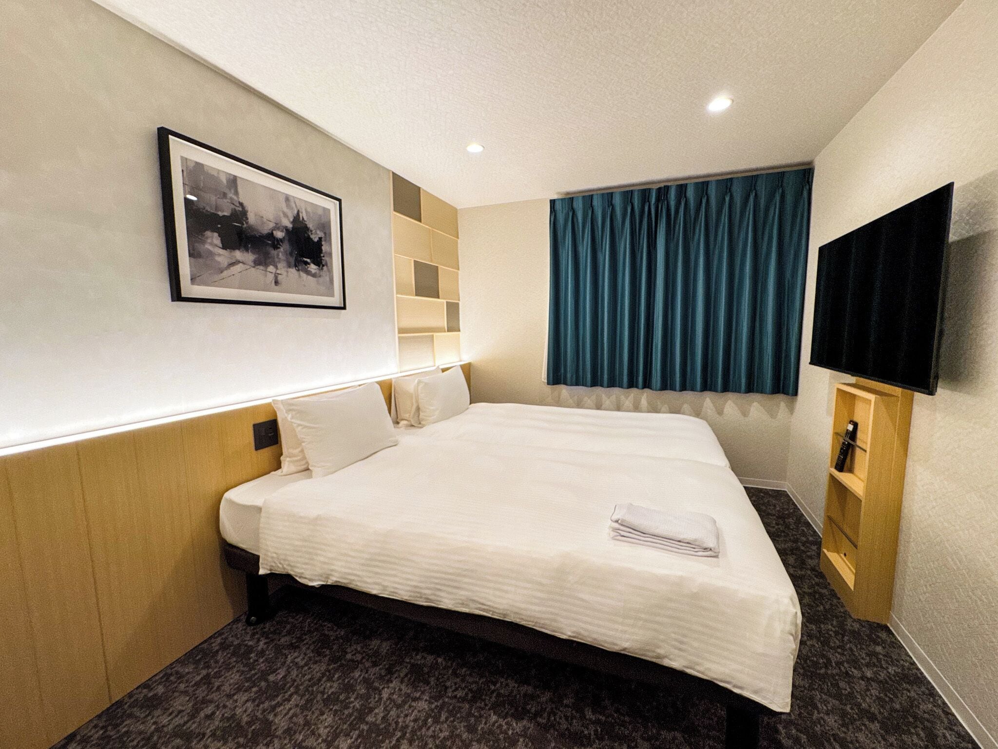 Quarto de hotel com uma cama grande, televisão montada, quadro na parede e janela com cortinas.