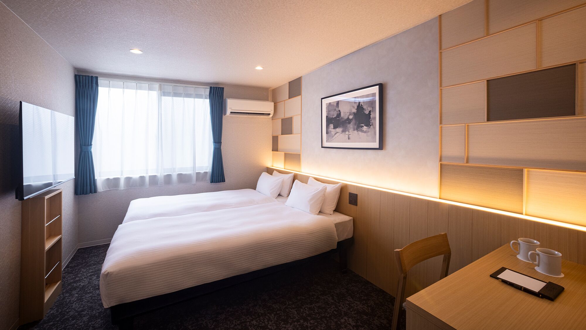 A hotel room with a large bed, a desk with two mugs, a chair, and a framed picture on the wall.