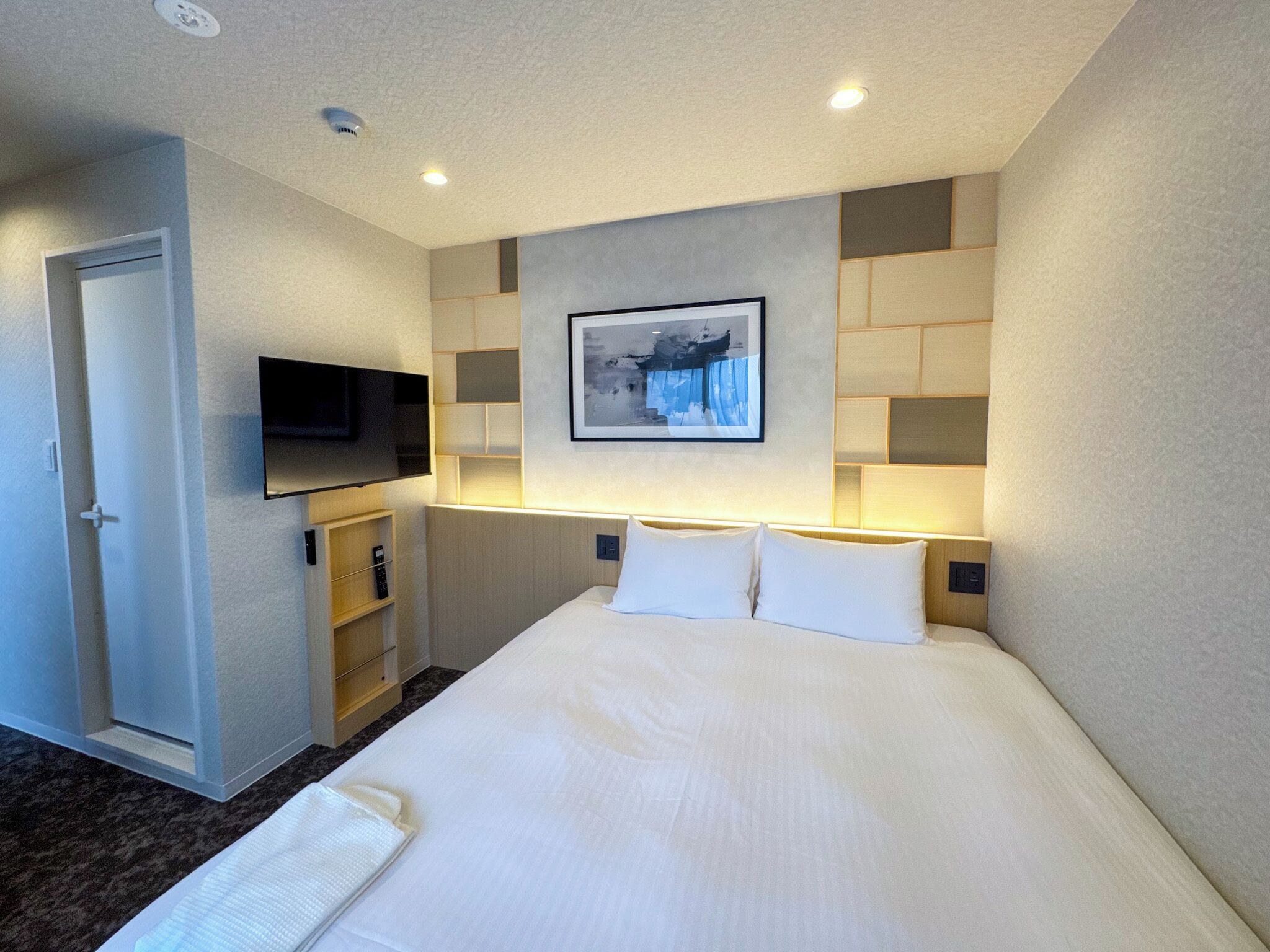 A hotel room with a large bed, a flat-screen TV mounted on the wall, a small seating area with a shelf, and a framed picture above the bed.