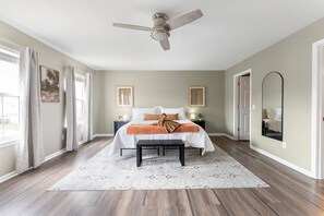 Quarto com cama, mesas de cabeceira, ventoinha no teto, espelho grande e janela com cortinas.