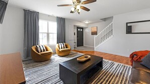 A modern living room with a ceiling fan, two armchairs, a coffee table, a staircase, and a large window.