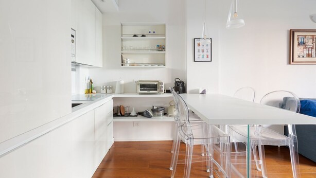 A modern kitchen with white cabinets, a white countertop, a built-in oven, and a dining area with transparent chairs.