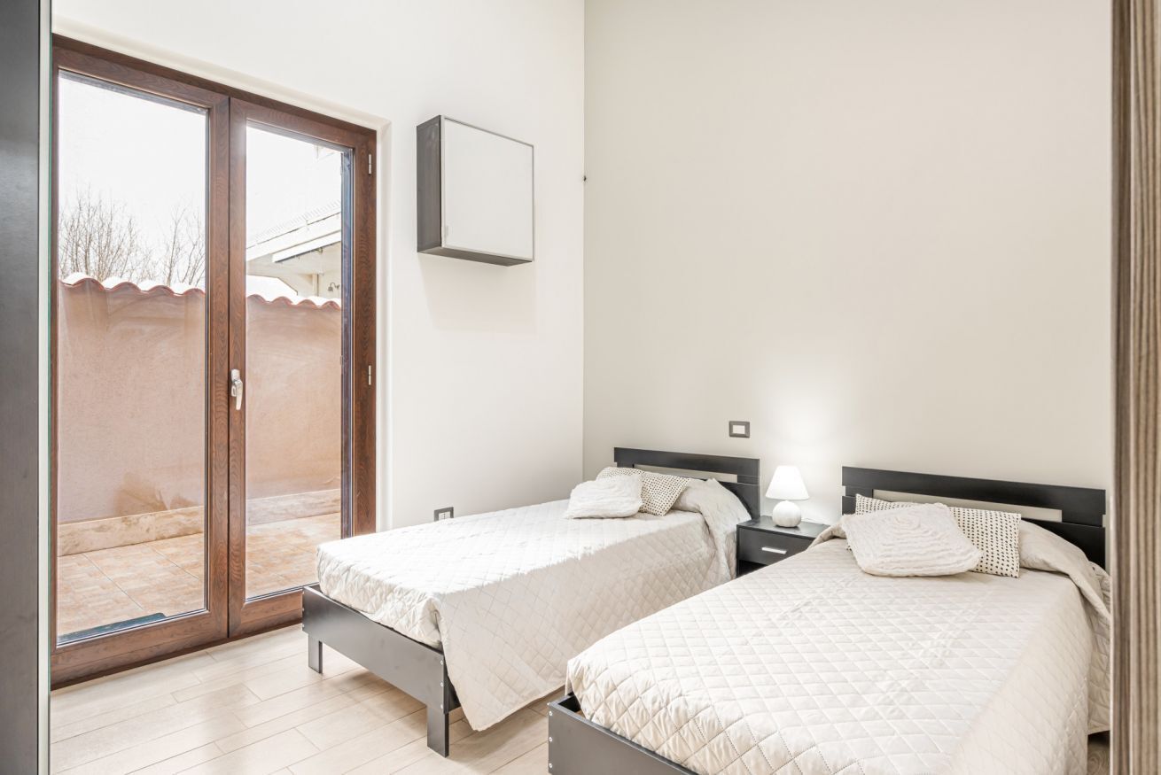 A bedroom with two beds, a sliding glass door to a balcony, and a bedside table with a lamp.
