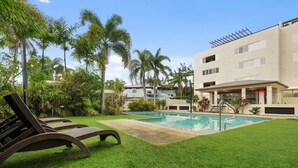 Un área de piscina con una tumbona, piscina y un edificio con múltiples ventanas y un porche cubierto.