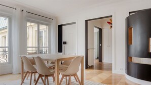 A modern dining area with a wooden table and chairs, large windows with curtains, and a view of the street.
