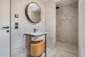 A bathroom with a marble wall, a round mirror, a sink with a towel rack, and a shower area. - Blueground | Le Marais, doorman & w/d, nr Bastille (Paris)