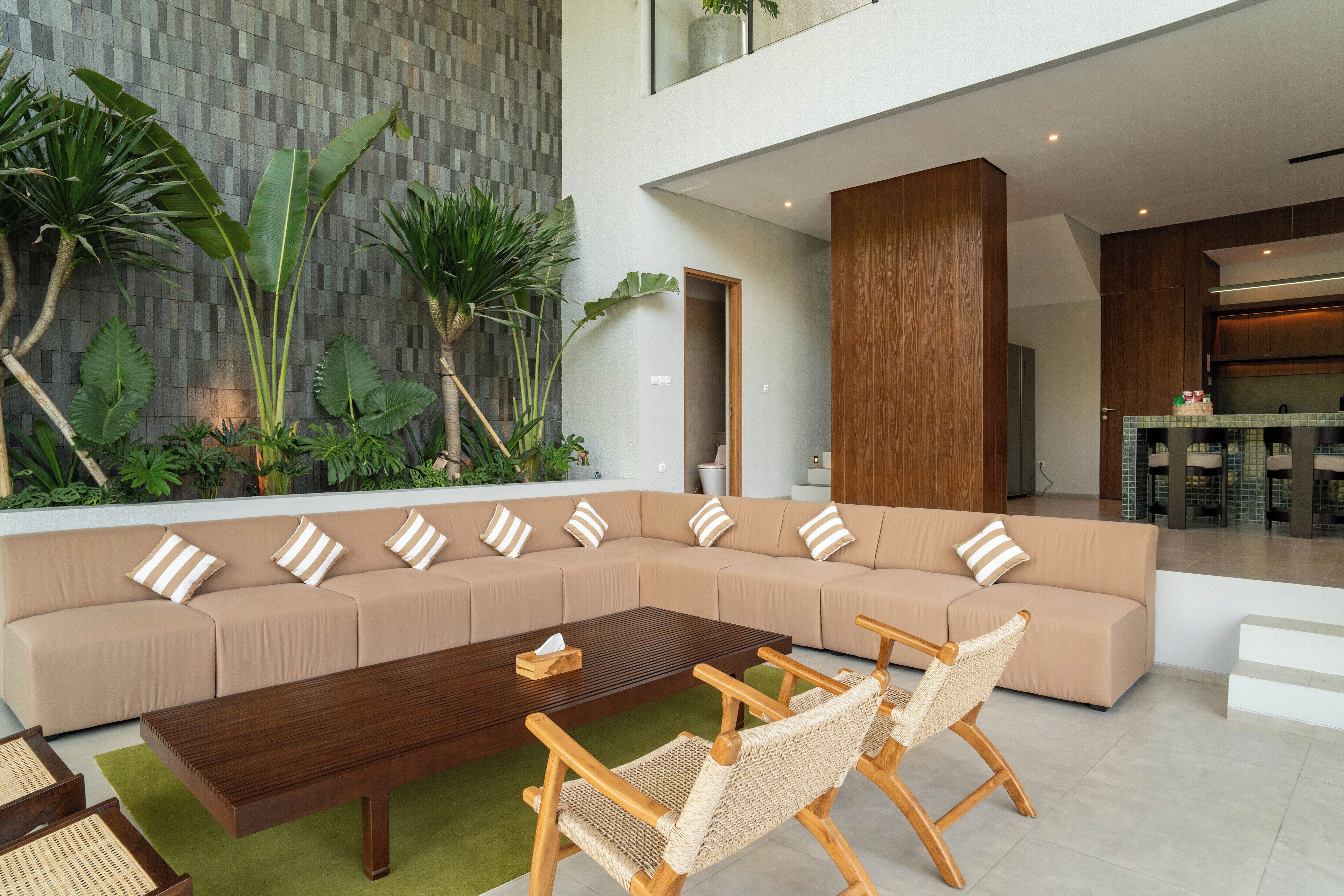 A modern living room with a large beige sectional sofa, a wooden coffee table, and wicker chairs. There is a green area rug, and the room has a view of a kitchen and a balcony with plants.