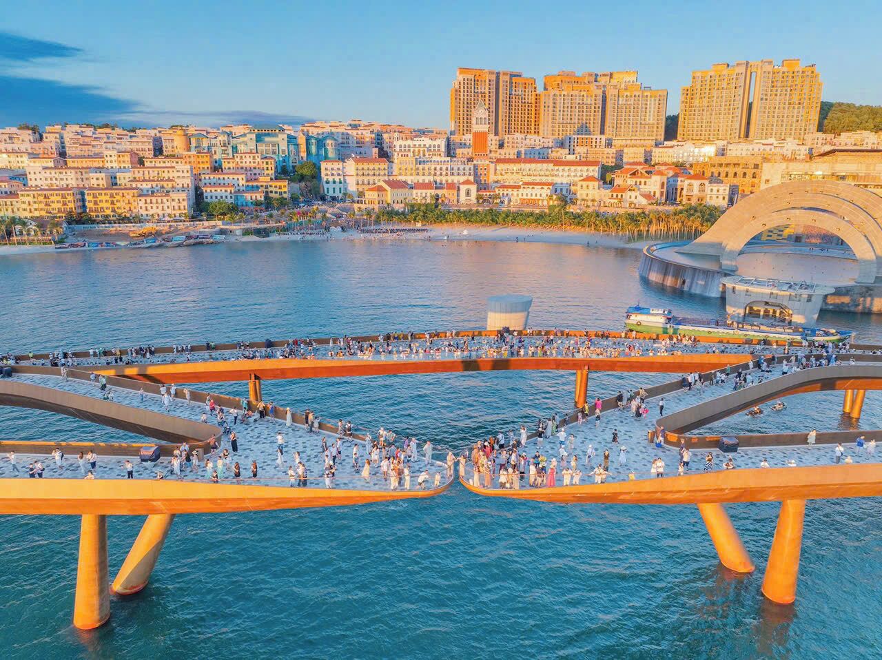 Un paseo marítimo con gente caminando, un puente y un paisaje urbano al fondo.